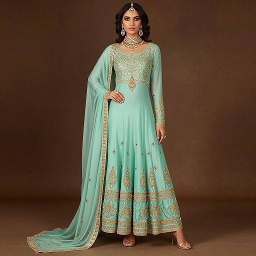 Photograph of a woman in a light turquoise traditional Indian lehenga with gold embroidery, wearing a matching choker and earrings, standing against a brown background