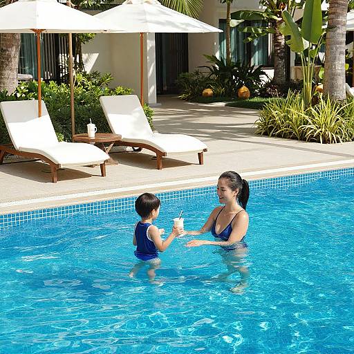 Photograph of a woman and young boy in a bright blue swimming pool, sharing a drink under white umbrellas, surrounded by sun loungers and tropical