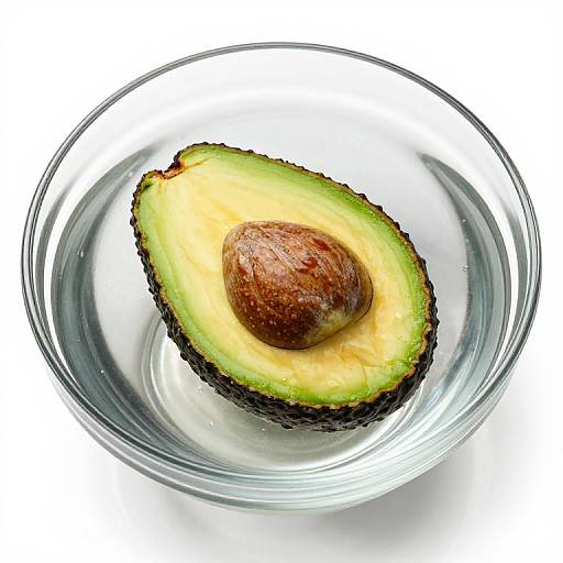 Photograph of a halved avocado with a brown seed, in a clear glass bowl, against a white background.