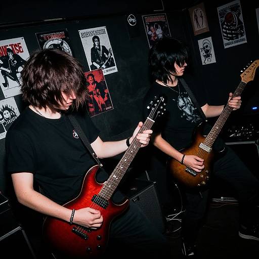 Photograph of two black-haired, pale-skinned male musicians with goth styles, playing red and brown electric guitars in a dark room with rock band