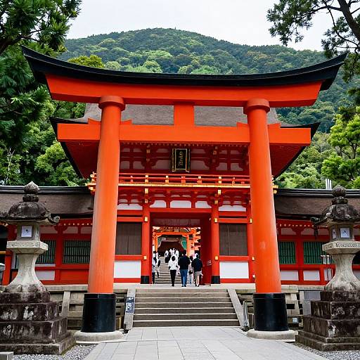 Photograph of a vibrant red torii gate with black accents, standing before a traditional Japanese shrine, surrounded by lush green trees and mountains in the background