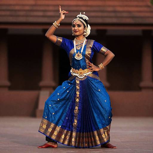 Photograph of a South Asian woman in traditional blue and gold dance costume, bejeweled, mid-dance pose, raised arm, stage background