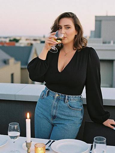 Photograph of a young woman with wavy brown hair, wearing a black V-neck blouse and high-waisted blue jeans, sipping wine on