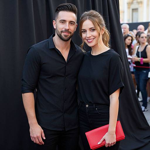 Photograph of a bearded, dark-haired man in a black shirt and a smiling woman with brown hair in a black top, holding a red clutch