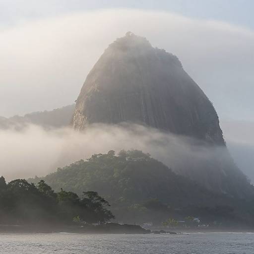 Photograph of a mist-covered, towering, green forested mountain with steep, rocky peak, partially obscured by fog, set against a calm lake.