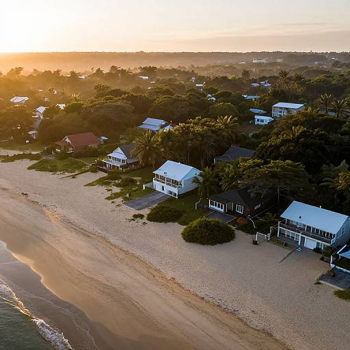Aerial photograph of a serene beach at sunset, featuring white and brown houses with blue roofs, surrounded by lush green trees and sandy shore. Warm golden