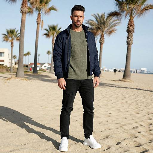 Photograph of a bearded man with short dark hair, wearing a navy jacket, green shirt, black jeans, and white sneakers, standing on a
