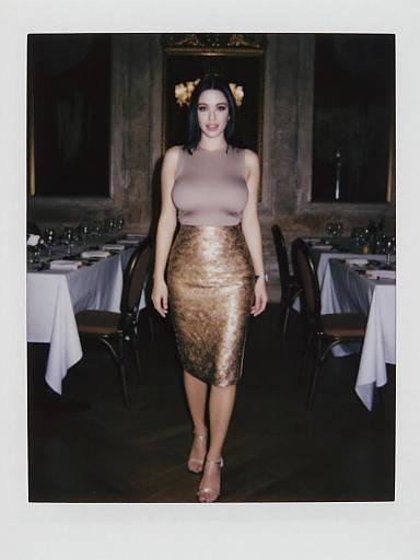 Photograph of a woman with pale skin, black hair, and large breasts, wearing a gold sequin, sleeveless, knee-length skirt, and