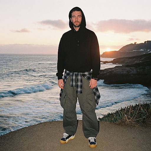 Photograph of a man with a beard, black hoodie, plaid shirt tied around waist, green pants, black shoes, standing on coastal path at