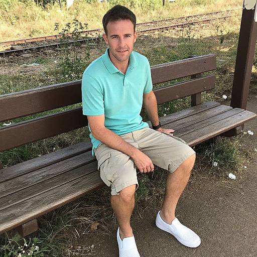 Photograph of a smiling, short-haired man in a light blue polo and beige shorts, sitting on a wooden bench in a sunny, grassy park