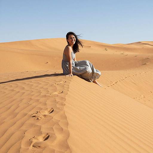 Photograph of a smiling Asian woman with long black hair, wearing a blue dress, sitting on a golden sand dune under a clear blue sky.