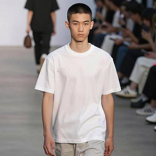 Photograph of a slim, young Asian male model with short black hair, wearing a plain white t-shirt and light gray pants, walking down a fashion