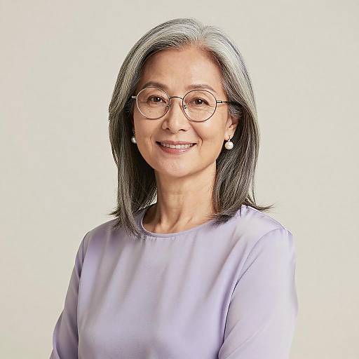 Photograph of an elderly Asian woman with gray hair, wearing glasses, pearl earrings, and a lavender blouse, smiling against a plain white background.
