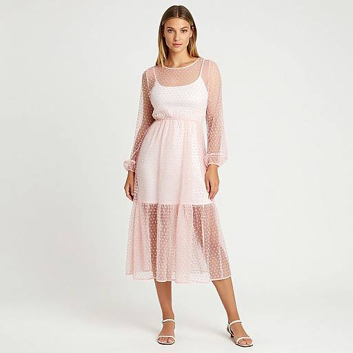 Photograph of a blonde woman in a white, sheer, lace, long-sleeve, knee-length dress and white sandals, standing against a plain