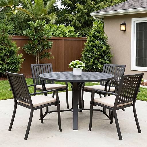 Photograph of a black patio set with four chairs and a round table, featuring a white flower pot, in a lush backyard with a wooden fence and