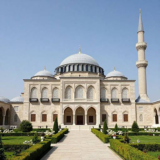 Photograph of a grand, beige Islamic mosque with silver domes, a tall minaret, and ornate arches, set in a well-man