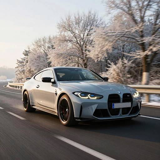 Photograph of a sleek, white BMW sports car with black wheels speeding down a snowy, tree-lined road at sunset.