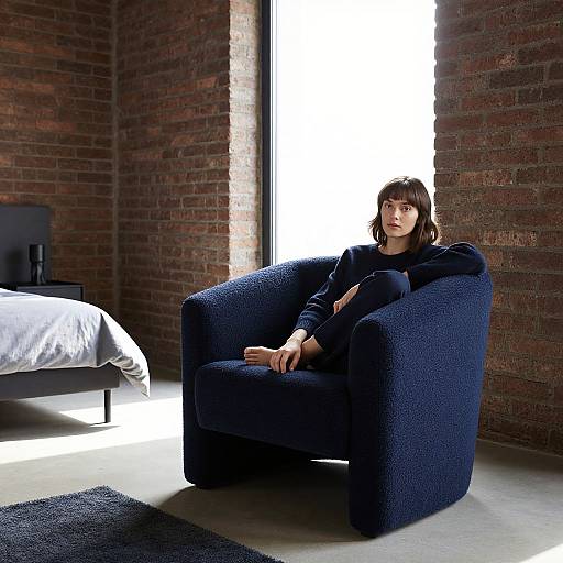 Photograph of a woman with short brown hair, wearing a navy sweater and black pants, sitting in a dark blue armchair against a brick wall,