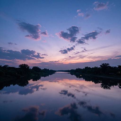 Photograph of a tranquil sunset over a still lake, with silhouetted trees on both sides, reflecting pink and blue sky with scattered clouds.