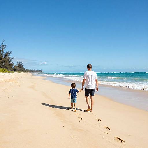 Photograph of a man in a white shirt and black shorts, and a toddler in a blue shirt and shorts, walking on a sunny, sandy beach