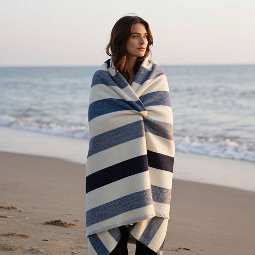 Photograph of a young woman with medium-length brown hair, standing on a beach, wrapped in a large, blue-and-white striped blanket, facing the