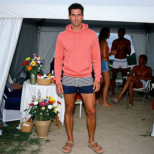 Photograph of a muscular man with short dark hair, wearing a coral hoodie, navy and white striped shorts, and sandals, standing in front of a