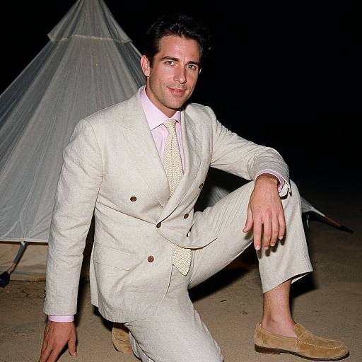 Photograph of a handsome man in a white double-breasted suit, pink shirt, and beige tie, posing confidently at night by a white tent.