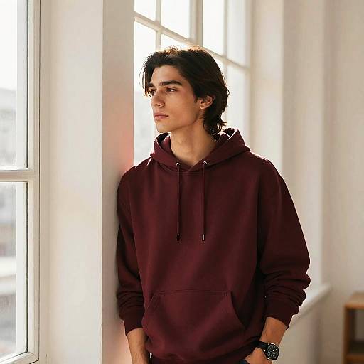 Photograph of a young man with medium-length dark hair, wearing a maroon hoodie, standing by a sunlit window, looking to the side.