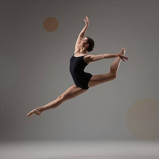 Photograph of a female ballerina mid-air in a black leotard, performing an elegant split jump against a gray background with subtle lens fl
