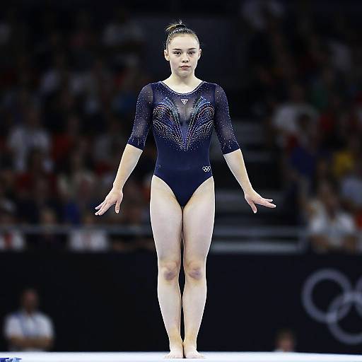 Photograph of a female gymnast in a dark blue, sparkly leotard with three-quarter sleeves, standing confidently on a gymnastics mat in