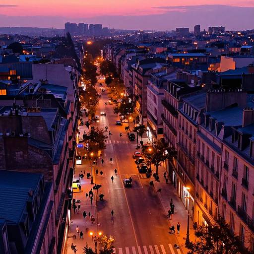 Aerial photograph of a vibrant city street at sunset, with orange streetlights, silhouetted pedestrians, and buildings lining the road.