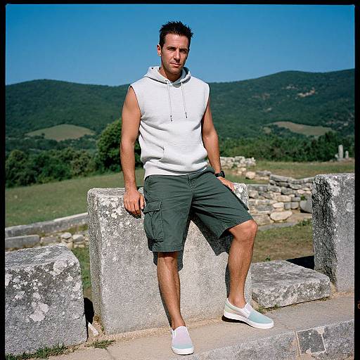 Photograph of a muscular man with short dark hair, wearing a white sleeveless hoodie, dark green cargo shorts, and white sneakers, sitting on ancient