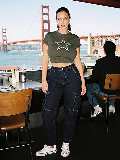 Photograph of a young woman with dark hair in a ponytail, wearing a green star crop top, black cargo pants, and white sneakers, standing