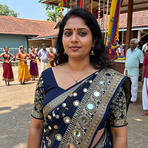 Photograph of a South Asian woman with dark hair in a black and gold embroidered sari, standing outdoors at a colorful festival.