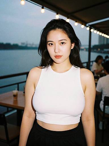 Photograph of an Asian woman with long black hair, wearing a white crop top and black pants, standing in a waterfront restaurant at dusk, with string