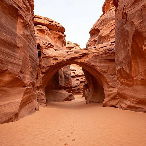 Photograph of a striking red sandstone archway in a narrow canyon, with smooth, layered rock walls and a bright white sky above. Desert sand