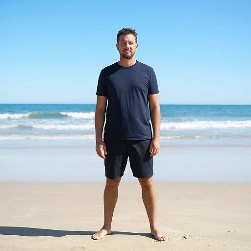 Photograph of a bearded man with short brown hair, wearing a black t-shirt and black shorts, standing barefoot on a sunny beach with blue