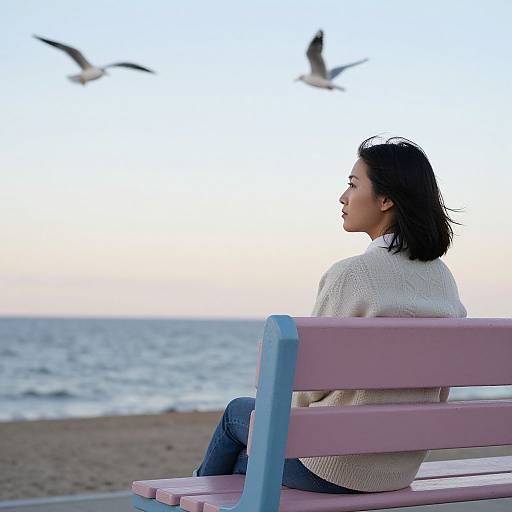 Photograph of an Asian woman with short black hair, wearing a white sweater and blue jeans, sitting on a pink bench, gazing at the ocean