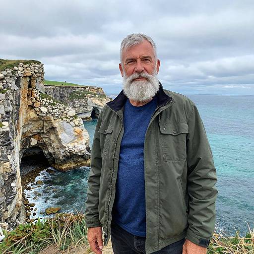 Photograph of an older man with a white beard, green jacket, and blue shirt, standing in front of a coastal cliff and ocean. Overcast