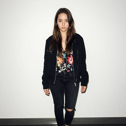 Photograph of a young woman with long brown hair, wearing a black jacket, colorful floral blouse, and ripped black jeans, standing against a plain white