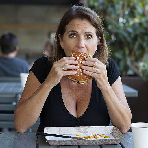 Photograph of a middle-aged woman with brown hair, wearing a black V-neck shirt, eating a sesame seed bagel at an outdoor café, with
