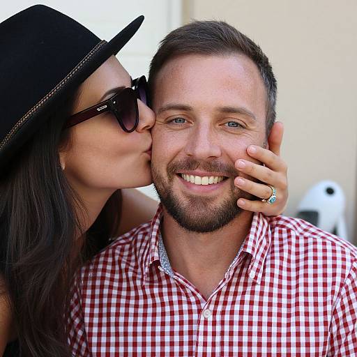 Photograph of a smiling bearded man with short brown hair, wearing a red checkered shirt, being kissed on the cheek by a woman with long