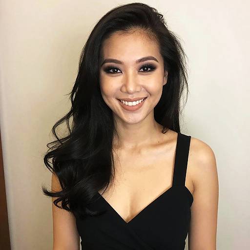 Photograph of a smiling Asian woman with long black wavy hair, wearing a black sleeveless dress, against a plain white background.
