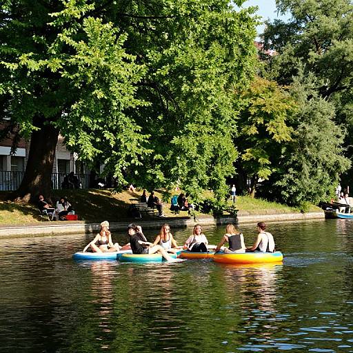 Photograph of six people in colorful inflatable boats floating on a calm, tree-lined lake, with a sunlit, shaded park background.