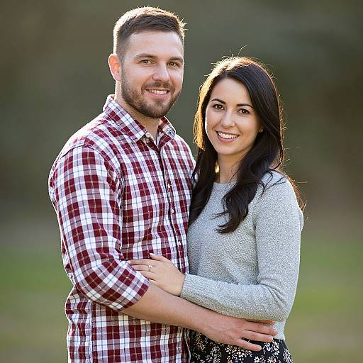 Photograph of a smiling couple; man with short brown hair, beard, red plaid shirt; woman with long black hair, gray sweater, standing