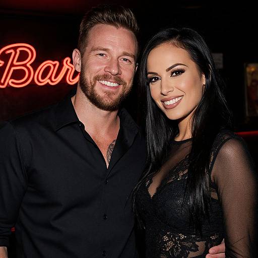 Photograph of a smiling bearded man in a black shirt and a smiling woman with long black hair in a lace dress, standing close together against a