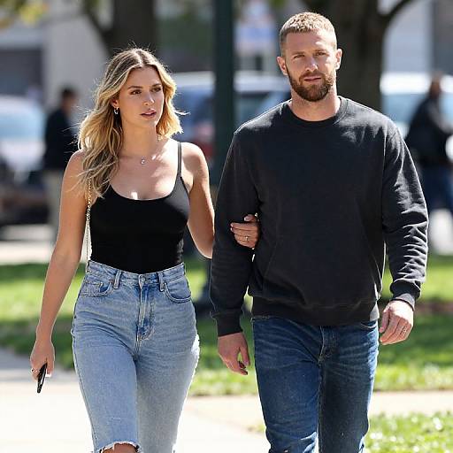Photograph of a blonde woman in a black tank top and high-waisted jeans, walking beside a bearded man in a black sweater and jeans
