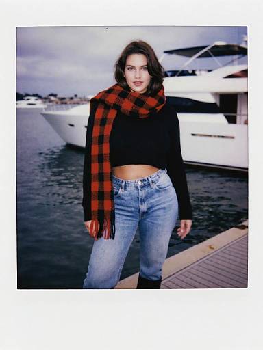 Photograph of a brunette woman with red and black plaid scarf, black crop top, and high-waisted jeans, standing by a marina