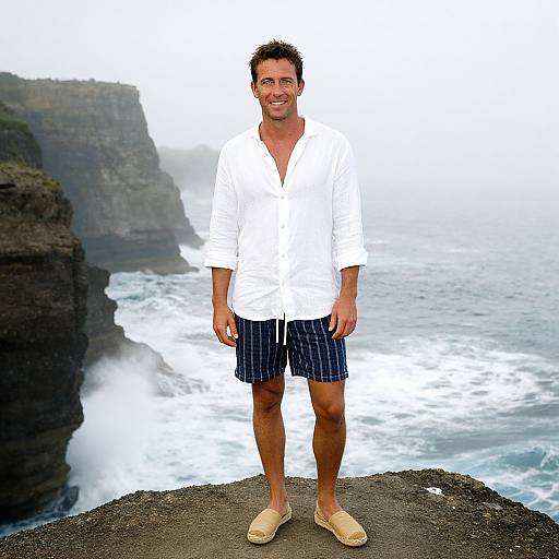 Photograph of a smiling man in a white shirt, blue striped shorts, and beige sandals, standing on a rocky cliff overlooking a misty ocean with