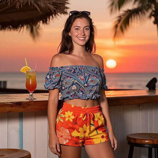 Photograph of a smiling young woman with long brown hair, wearing an off-shoulder blue patterned top and orange floral shorts, standing at a